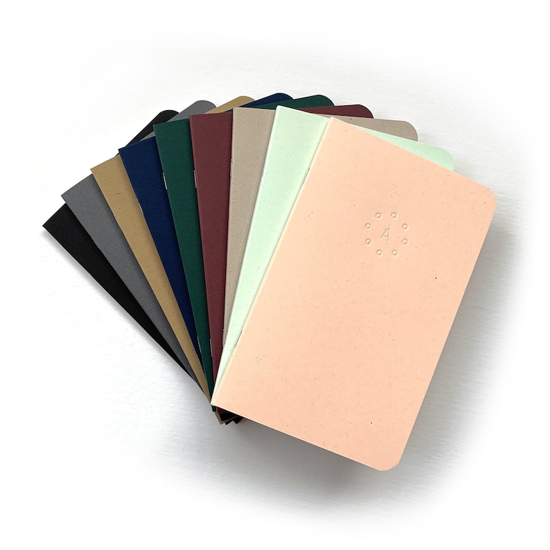 K&ouml;nnte beinhalten: Ein Stapel aus zehn verschiedenen farbigen Notizb&uuml;chern mit abgerundeten Ecken. Das oberste Notizbuch ist pfirsichfarben mit einem kleinen Kreisdesign und dem Buchstaben "A" in der Mitte.