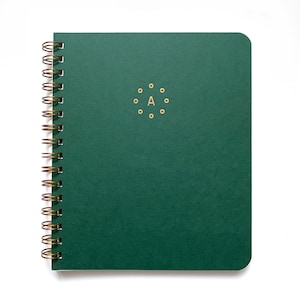 May include: A dark green spiral-bound notebook with a gold-colored spiral and the letter "A" surrounded by gold dots. The notebook has a rounded corner and is set against a white background.