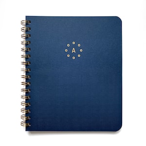 May include: A navy blue spiral-bound notebook with a gold spiral binding. The cover features the letter "A" surrounded by gold dots. The notebook has a textured cover and rounded corners, perfect for journaling or note-taking.