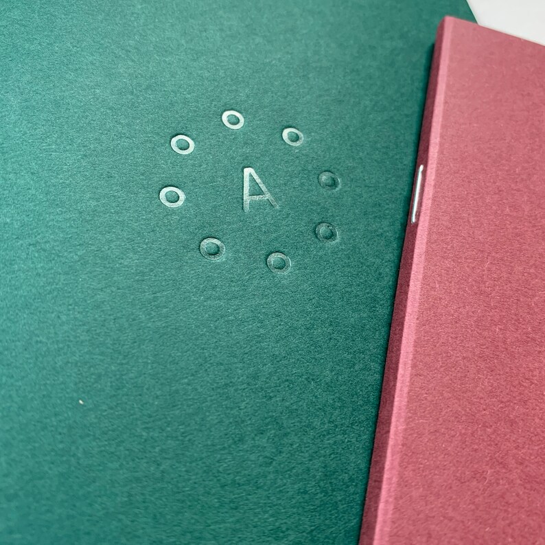 K&ouml;nnte beinhalten: Zwei Notizb&uuml;cher, eines gr&uuml;n und eines burgunderrot, mit einem silbernen, gepr&auml;gten Buchstaben "A" umgeben von einem Kreis aus Punkten auf dem gr&uuml;nen Notizbuch.
