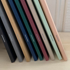 K&ouml;nnte beinhalten: Ein Satz von zehn Notizb&uuml;chern mit verschiedenen farbigen Umschl&auml;gen, jedes mit einer silbernen Naht entlang des R&uuml;ckens.