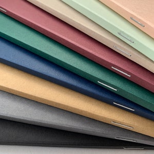 K&ouml;nnte beinhalten: Ein Stapel von acht Notizb&uuml;chern mit verschiedenen farbigen Umschl&auml;gen, darunter Schwarz, Grau, Beige, Blau, Gr&uuml;n, Burgunder, Hellbraun und Hellgr&uuml;n. Die Notizb&uuml;cher sind mit einem Stich entlang des R&uuml;ckens gebunden.