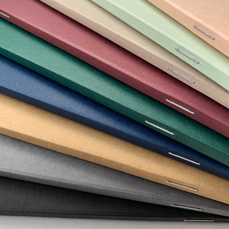 K&ouml;nnte beinhalten: Ein Stapel von acht Notizb&uuml;chern mit verschiedenen farbigen Umschl&auml;gen, darunter Schwarz, Grau, Beige, Blau, Gr&uuml;n, Burgunder, Hellbraun und Hellgr&uuml;n. Die Notizb&uuml;cher sind mit einem Stich entlang des R&uuml;ckens gebunden.