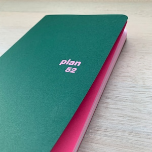 Peut inclure: Un carnet à couverture rigide vert avec le texte "plan 52" imprimé en rose sur la couverture.