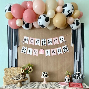 May include: A pink and black cow-themed birthday party backdrop with a banner that reads "Moo Moo I'm Two". The backdrop is decorated with a garland of balloons in pink, beige, and black with a cow print. A table with a black and white striped tablecloth is set up in front of the backdrop. The table is decorated with a cake, hay bales, and other party decorations.