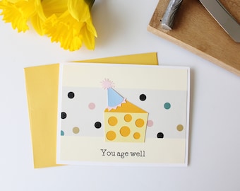 May include: A yellow and white birthday card with a cheese wedge wearing a party hat. The card has a gray background with black, gold, pink, and teal polka dots. The text on the card reads "You age well."
