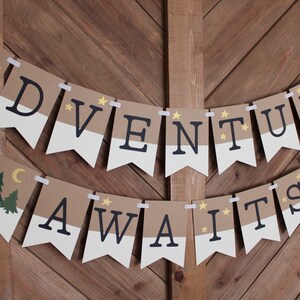Puede incluir: Una guirnalda con las palabras "ADVENTURE AWAITS" en letras negras sobre banderines marrones y blancos. La guirnalda incluye ilustraciones de &aacute;rboles, una luna creciente y estrellas. Est&aacute; colgada con cinta blanca en una puerta de madera.