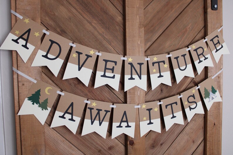 Puede incluir: Una guirnalda con las palabras "ADVENTURE AWAITS" en letras negras sobre banderines marrones y blancos. La guirnalda incluye ilustraciones de &aacute;rboles, una luna creciente y estrellas. Est&aacute; colgada con cinta blanca en una puerta de madera.
