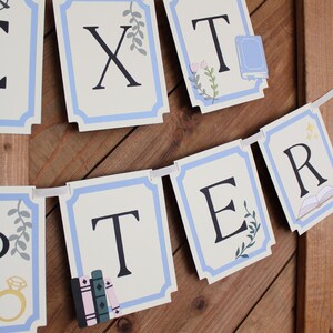 May include: A banner with individual rectangular cards spelling out "XTER" in black letters. Each card has a light blue border and decorative elements like books, flowers, and a ring. The banner is strung on a white ribbon against a wooden background.