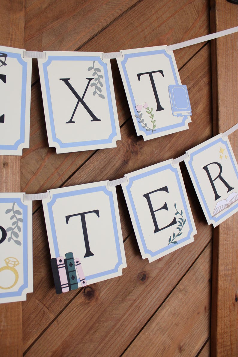 May include: A banner with individual rectangular cards spelling out "XTER" in black letters. Each card has a light blue border and decorative elements like books, flowers, and a ring. The banner is strung on a white ribbon against a wooden background.