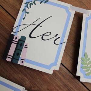 May include: A banner with the word "Her" in elegant script, framed by a light blue border and decorative leaves. The banner includes illustrations of books in pink, green, and black. The banner is strung with a white ribbon.