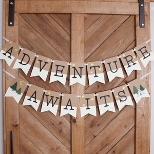 Puede incluir: Un cartel con las palabras "ADVENTURE AWAITS" en letras negras sobre un dise&ntilde;o de bander&iacute;n marr&oacute;n y blanco. El cartel est&aacute; decorado con estrellas doradas, &aacute;rboles y una luna creciente. Cuelga de una puerta de madera.