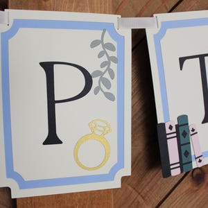 May include: A banner with the letter "P" in black, a gold ring, and gray leaves. The banner has a light blue border and is strung with white ribbon. The background is a wooden surface.