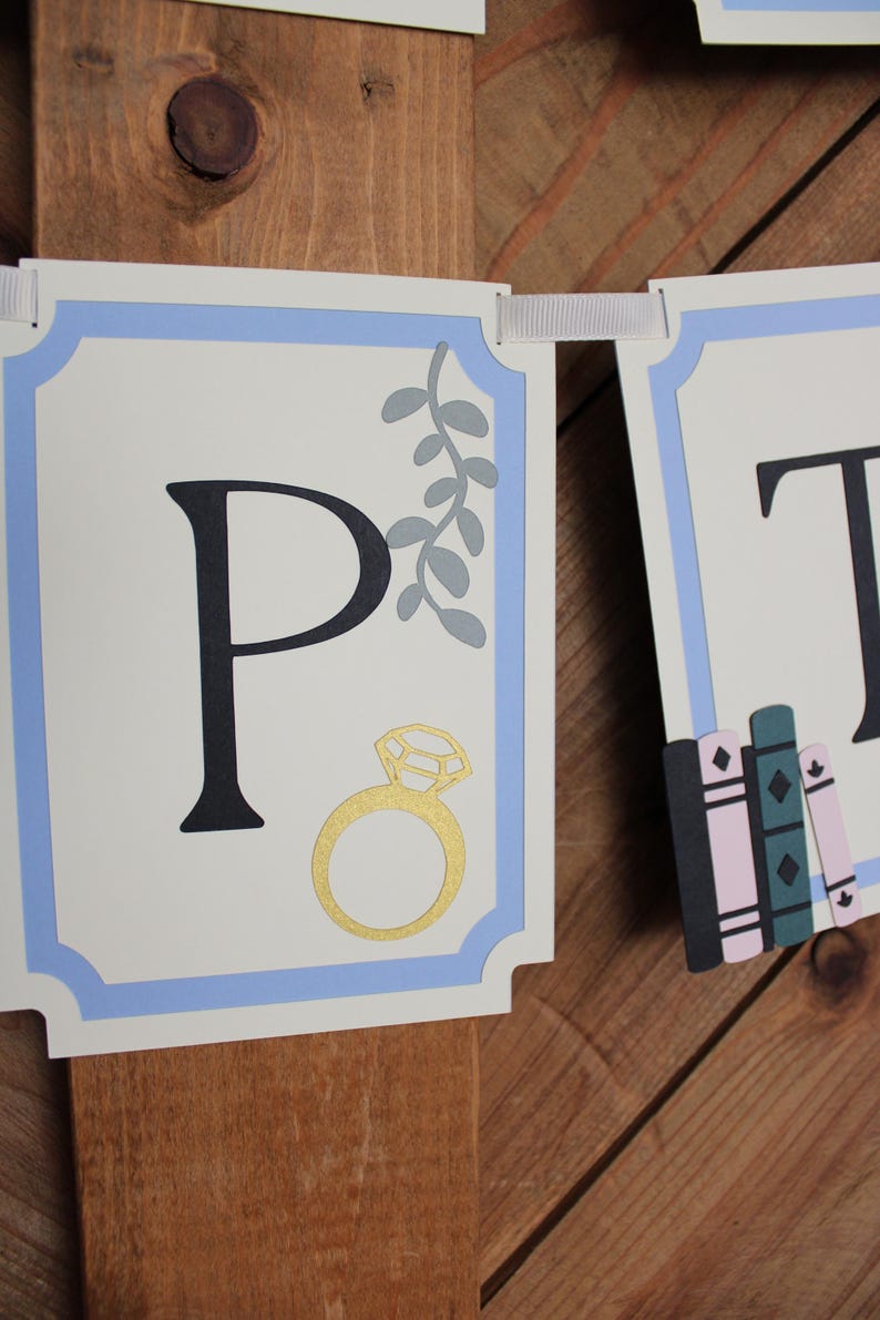 May include: A banner with the letter "P" in black, a gold ring, and gray leaves. The banner has a light blue border and is strung with white ribbon. The background is a wooden surface.
