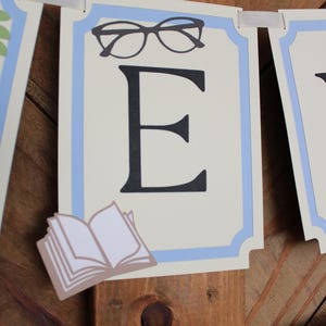 May include: A banner with a light blue border and a cream-colored background. The banner features a large black letter "E", a pair of glasses, and a small open book. The banner is strung with a white ribbon.