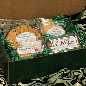 Può includere: Una scatola regalo verde piena di dolcetti a tema irlandese, tra cui un pane soda irlandese dolce, uno scone dolce al burro, una fetta di torta al whisky e caffè irlandese, caramelle artigianali e tè.