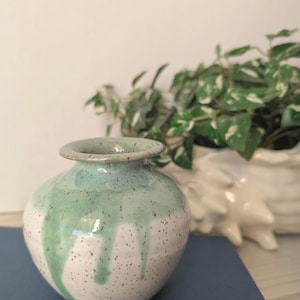 May include: A small, handcrafted ceramic vase with a rounded body and a narrow neck. The vase is primarily white with speckled detailing and a turquoise-green glaze that drips down the sides. A potted plant is in the background.
