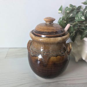 May include: A ceramic sugar bowl with a lid and small handles. The bowl is a mix of brown and tan hues, with a glossy finish. A small, light pink spoon rests on the rim. The bowl is likely handmade and adds a rustic touch to any kitchen.