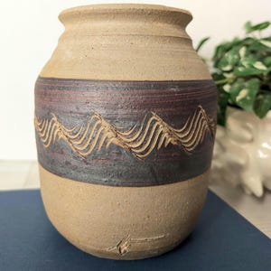 May include: A handcrafted ceramic vase with a beige and dark brown color scheme. The vase features a textured, wave-like design carved into the dark brown band. The vase has a rounded base and a slightly flared rim, showcasing a rustic aesthetic.