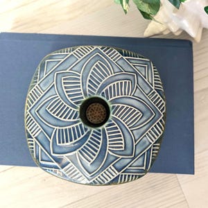 May include: A square, blue ceramic flower frog with a central hole and a decorative, geometric pattern in white. The flower frog is designed to hold flowers in place. The design is reminiscent of a mandala.
