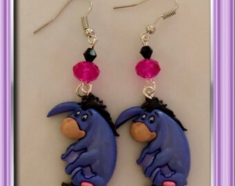 May include: A pair of dangle earrings featuring the character Eeyore from Winnie the Pooh. The earrings have silver-tone hooks, black and pink beads, and a purple Eeyore charm with a pink tail. The text "Stacyleerainbows" is at the bottom.
