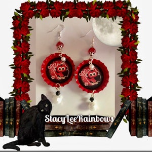 May include: Red and black owl bottle cap earrings with a white pearl and black bead accents. The earrings are hanging from silver hooks.