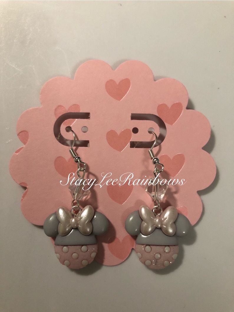 May include: A pair of grey and pink Minnie Mouse earrings with a white bow and clear crystal accents. The earrings are on a pink heart-shaped display card with the text "Stacy Lee Rainbows".