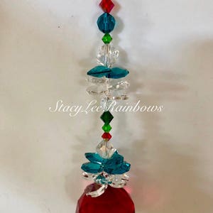 Op de afbeelding: Een rode kristallen zonenvanger met turquoise, groene en heldere kristallen accenten. De zonenvanger hangt aan een zilveren ketting.