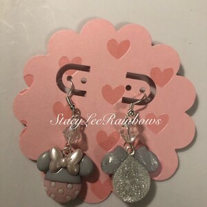 May include: A pair of dangle earrings featuring a grey Minnie Mouse head with pink polka dots and a silver bow on one side and a silver glitter ball on the other side. The earrings are hanging from silver hooks on a pink background with hearts.