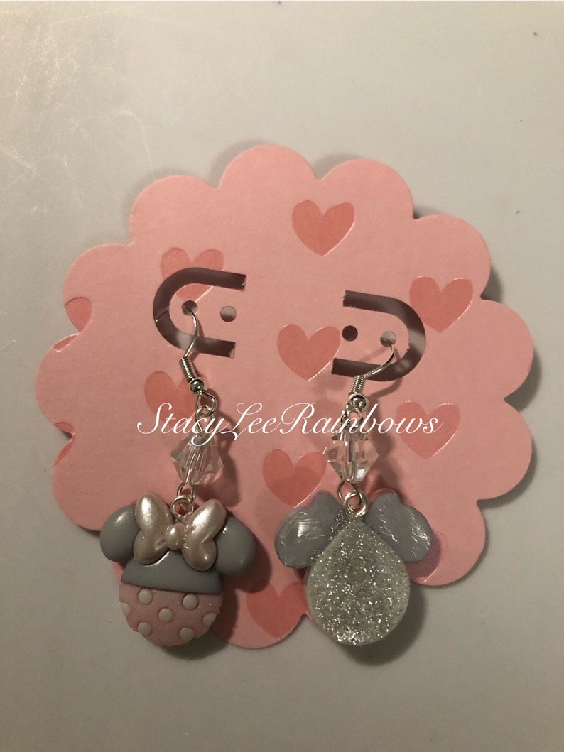 May include: A pair of dangle earrings featuring a grey Minnie Mouse head with pink polka dots and a silver bow on one side and a silver glitter ball on the other side. The earrings are hanging from silver hooks on a pink background with hearts.