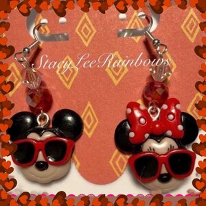 May include: A pair of dangle earrings featuring a red and black Mickey Mouse and Minnie Mouse design. The earrings have red and clear crystal beads and silver hooks. The earrings are on a pink and yellow background with the text "Stacy Lee Rainbows".