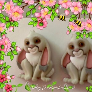 May include: A pair of white bunny rabbit earrings with pink ears and gray fur. The earrings are surrounded by pink flowers and bees.