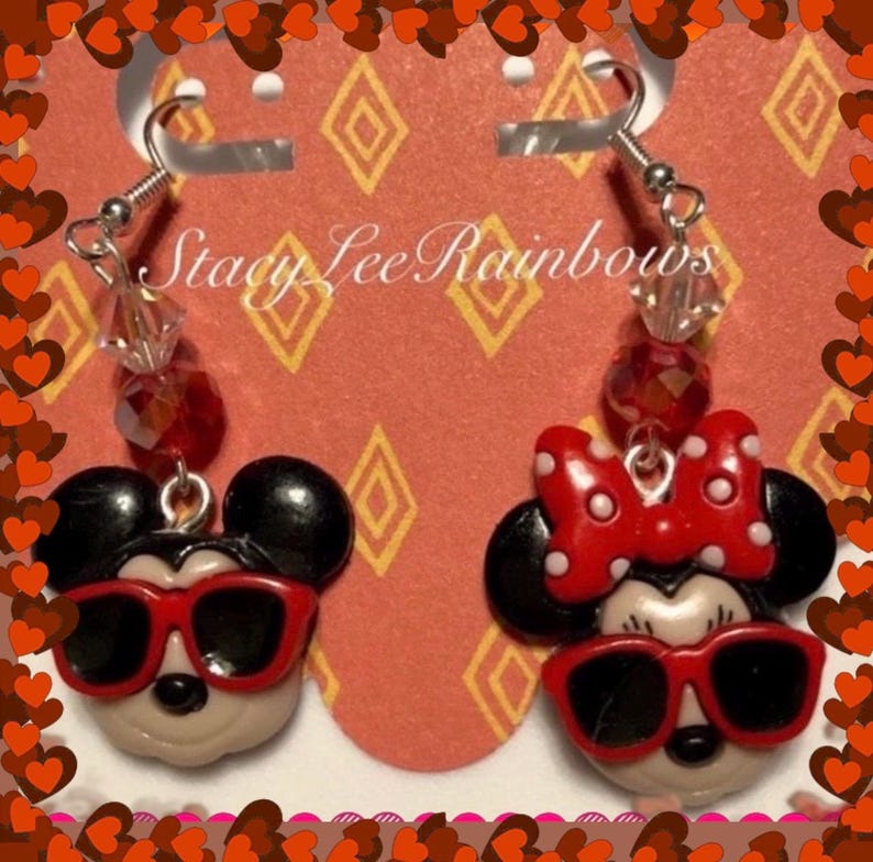 May include: A pair of dangle earrings featuring Mickey Mouse and Minnie Mouse wearing sunglasses. The earrings are made of polymer clay and have red and black accents. The earrings are hanging from silver hooks with red and clear beads.
