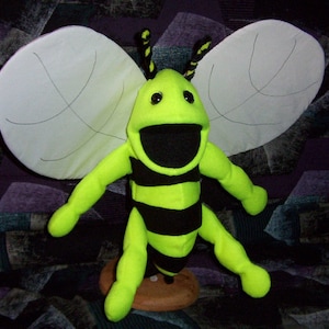 May include: A bright yellow and black bee puppet with large white wings. The puppet has a large, open mouth and black eyes.