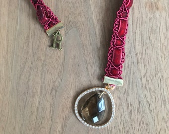 May include: A burgundy red braided leather necklace with a gold pendant featuring a teardrop-shaped brown gemstone surrounded by clear rhinestones.