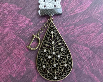 May include: A white lace bookmark with a metal charm attached. The charm is a teardrop shape with a filigree design and clear rhinestones. The letter "D" is attached to the charm.