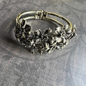 May include: A silver bangle bracelet with a floral design. The bracelet is made of two bands of metal that are connected by a hinge. The flowers are arranged in a cluster around the bracelet.