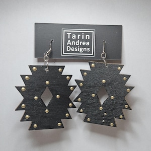 Puede incluir: Un par de pendientes de madera negra con detalles dorados en forma de estrella geométrica. Los pendientes cuelgan de ganchos plateados. Los pendientes están etiquetados como "Tarin Andrea Designs".