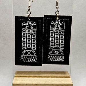 May include: A pair of black dangle earrings with a silver hook. The earrings feature a white design of a stylized eagle with a crown and a star.