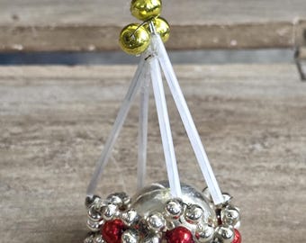 May include: A vintage Christmas ornament featuring a silver glass ball with red and silver bead accents. The ornament is suspended by clear plastic supports and topped with three gold-colored beads.