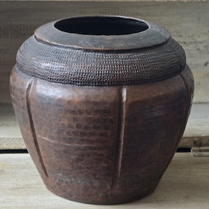 May include: A dark brown, rounded metal vessel with a wide opening and a thick, textured rim featuring a cross-hatched pattern. The body of the container has a hammered finish with vertical indentations that create a paneled appearance.