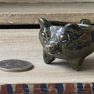 May include: A small, dark green ceramic pig planter with a glossy finish. The pig has a detailed face and is resting on three small legs. A quarter is next to the planter for size comparison. The planter is on a wooden surface.