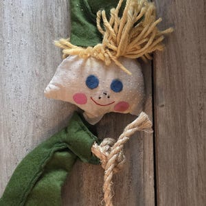 May include: A whimsical handmade elf doll with a green felt hat and outfit, a white dotted face with blue eyes and pink cheeks, and yellow yarn hair. The doll is holding a rope and is attached to a wooden surface.