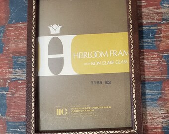 May include: A brown picture frame with a gold trim and a label that reads "Heirloom Frame with Non Glare Glass" and "1165 E3". The label also includes a logo for Intercraft Industries Corporation.