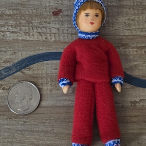 May include: A small doll wearing a red knit outfit with blue and white trim. The doll has a matching blue and white knit hat. The doll's face is painted with rosy cheeks and red lips. A U.S. quarter is next to the doll.