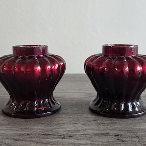 May include: Two antique, ruby red glass candle holders. Each has a fluted, bulbous body and a flared base. The candle holders are displayed on a weathered, gray wooden surface. The glass has a glossy finish.
