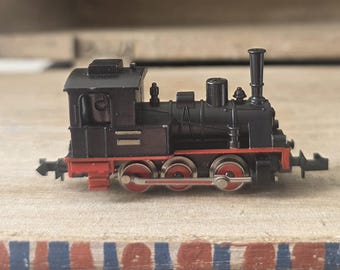 Op de afbeelding: Een zwarte miniatuur stoomlocomotief met een opvallende schoorsteen, een open cabine en een rood chassis. De trein heeft drie wielstellen met rode centra en zilverkleurige metalen drijfstangen aan beide zijkanten van het model.