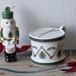 May include: Two Christmas ornaments: a ceramic nutcracker soldier and a drum. The nutcracker is white with green and gold accents, a red ribbon, and a gold string. The drum is white with green trim, a gold string, and a red ribbon.