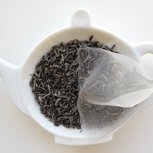 May include: A white ceramic teapot-shaped dish filled with loose green tea leaves and a tea bag. The tea leaves are a dark green color, and the tea bag is a light, translucent material. The dish has a handle and a spout.