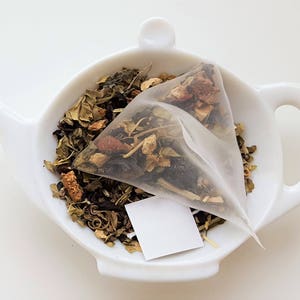 May include: A white ceramic teapot with a handle and a spout, filled with a tea bag containing a blend of green tea, dried fruit, and herbs.